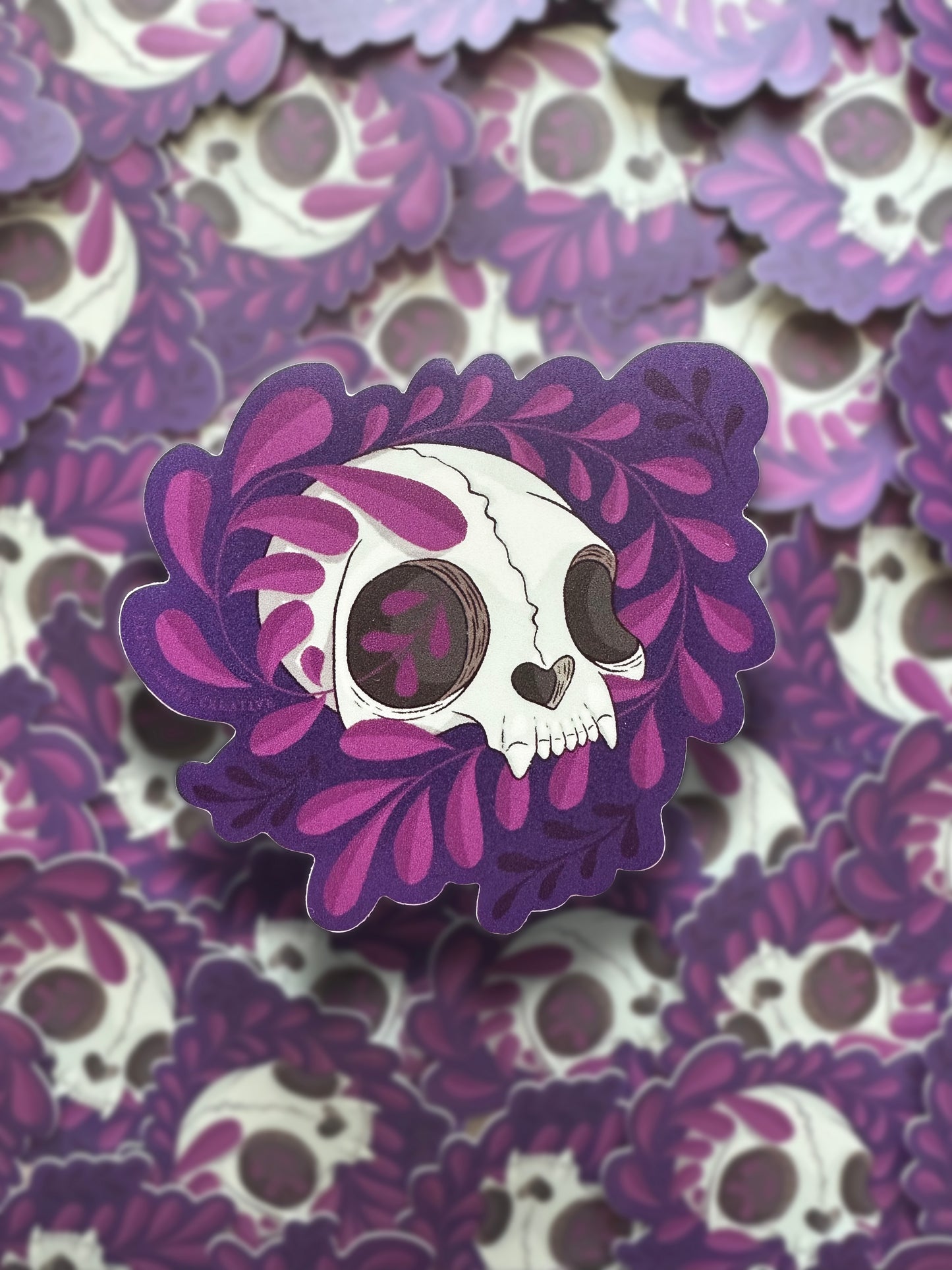 A sticker with a die-cut design featuring a cat skull with purple vines around it, against a repeating background of the same sticker.