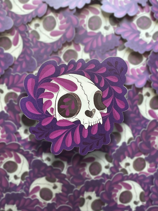A sticker with a die-cut design featuring a cat skull with purple vines around it, against a repeating background of the same sticker.