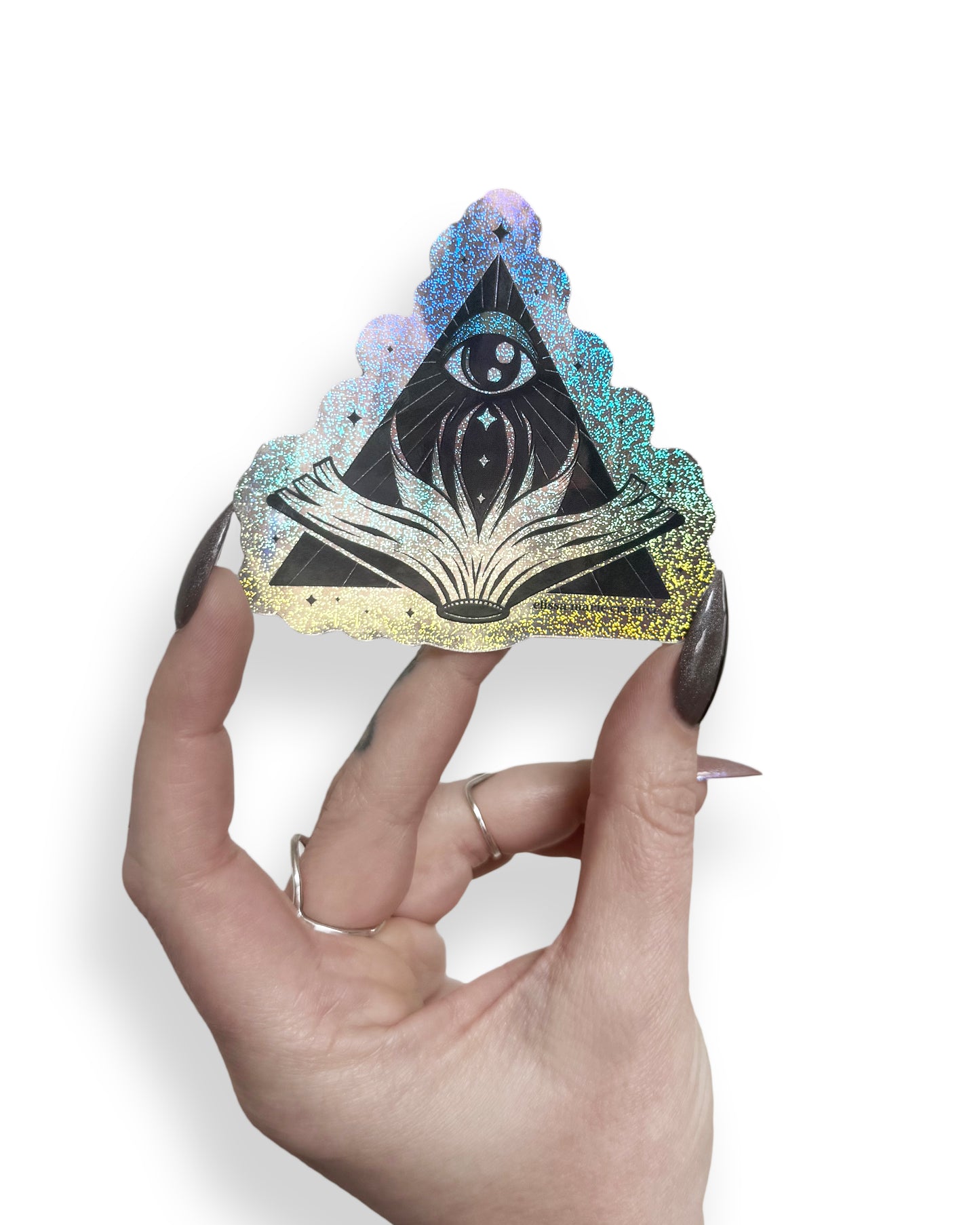 A rainbow-glitter sticker with a triangular design featuring an eye in the top center and an open book below, behind held by a hand with long witchy nails, on a white background.