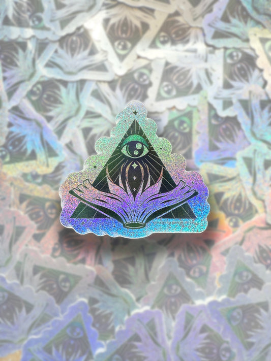 A multicolored glitter sticker with a triangular design, featuring an eye in the top center and an open book below it, displayed on a surface with a blurred background.