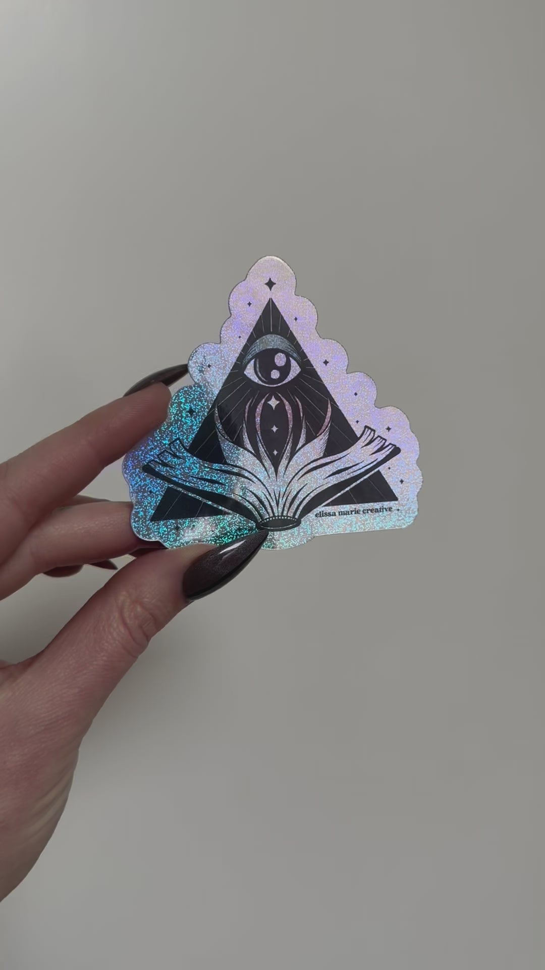 A holographic glitter sticker of a triangular design featuring an eye in the top center and an open book below it, being held and rotated by a hand with witchy nails, on a light grey background.
