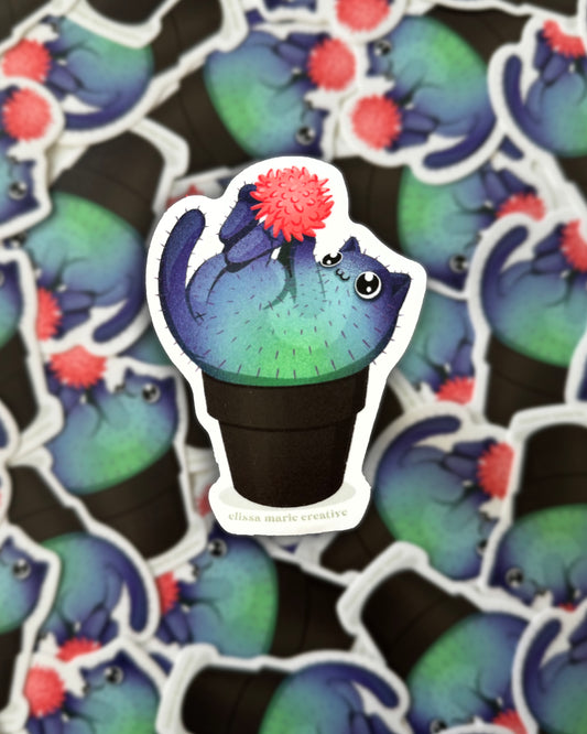 A die-cut vinyl sticker of an illustration featuring a green and purple cat-shaped cactus with a coral colored blossom, in a black pot. The sticker is displayed over a blurred background.