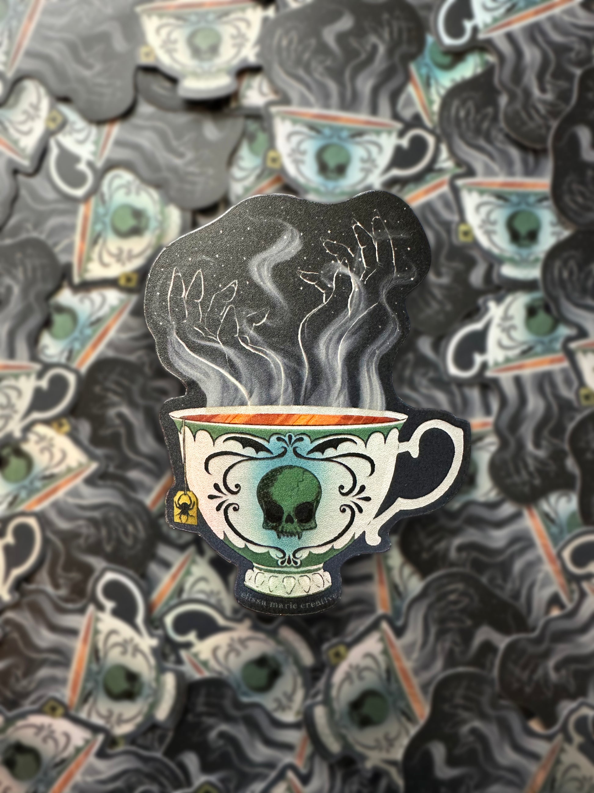 A die-cut sticker with an illustrated design of a teacup with a vampire skull painted on it, and hand-shaped steam rising from the tea. The sticker is displayed on top of a repeating background of the same sticker.