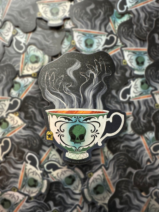 A die-cut sticker with an illustrated design of a teacup with a vampire skull painted on it, and hand-shaped steam rising from the tea. The sticker is displayed on top of a repeating background of the same sticker.